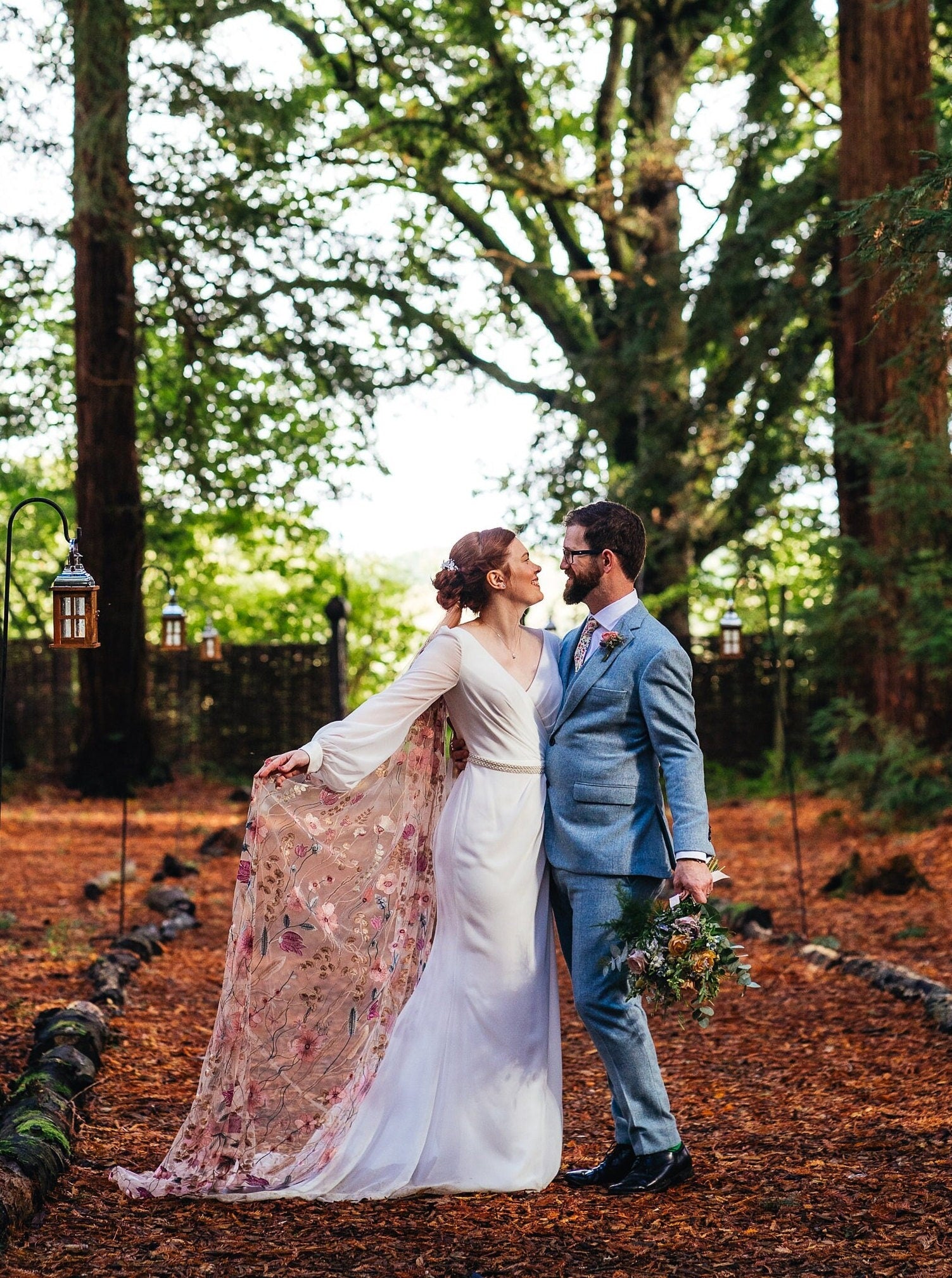 Bride and groom embracing in woodlands. Bride wears an elegant wedding dress and one of my veils called Dawn. Dawn is made from super soft dusky pink tulle, embroidered with flowers and leaves in cream, russet, pink, and teal.
